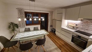 Et moderne køkken med indbygget ovn, et spiseområde med et glasbord og fire stole, samt et vindue med gardiner.