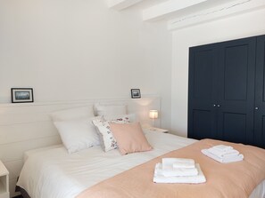 Un lit bien fait, avec du linge de lit blanc et un plaid de couleur pêche, une table de chevet avec une lampe et une porte bleu foncé.