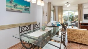 A modern dining area with a glass table and chairs, a painting of a beach scene, and a view of the outdoors through a sliding door.