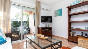 A living room with a glass coffee table, a television on a wooden stand, a bookshelf, and a sliding glass door leading to an outdoor area.