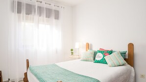 Un dormitorio con cama, cabecera de madera, mesita de noche con lámpara y ventana con cortinas traslúcidas.
