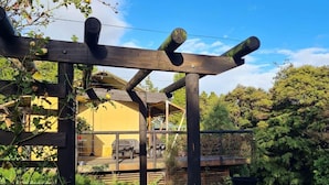 Una pérgola de madera con diseño rústico, un camino de ladrillo y una zona verde con un edificio amarillo al fondo.