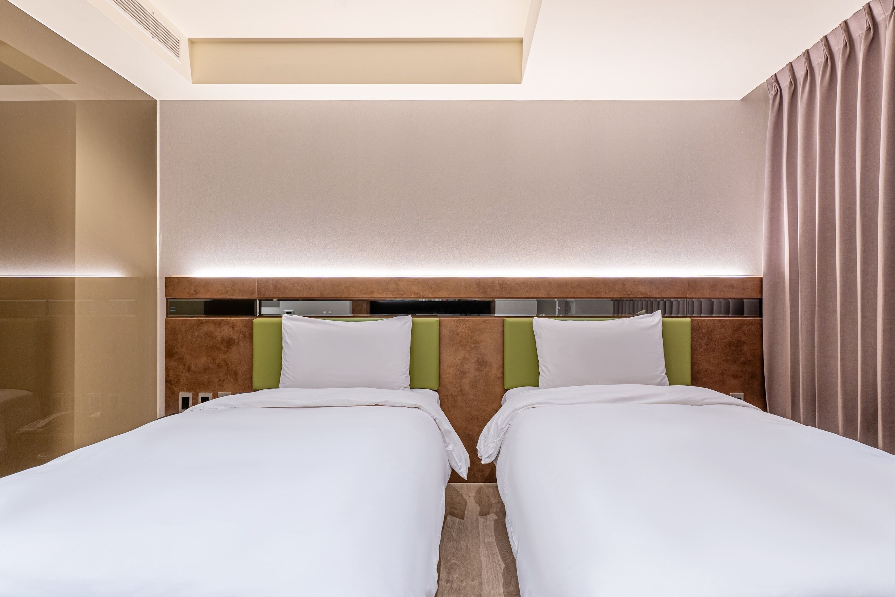 A hotel room with two beds, a headboard with a light fixture, and a large window with curtains.