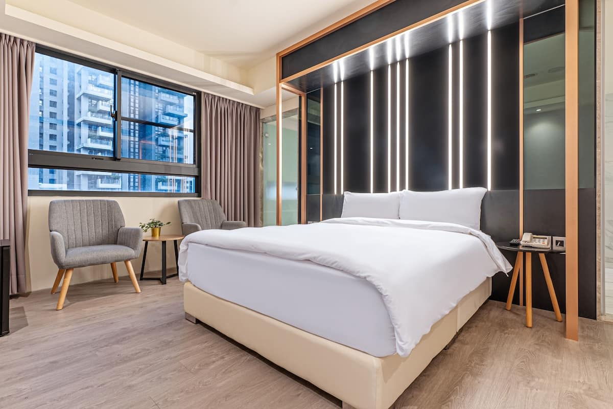 A modern hotel room with a large bed, a seating area with a chair and table, a small side table, and a view of the city through large windows.