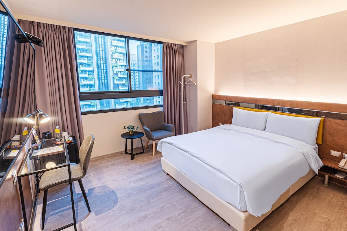 A hotel room with a large bed, a desk with a chair, a small table, and a window with curtains.