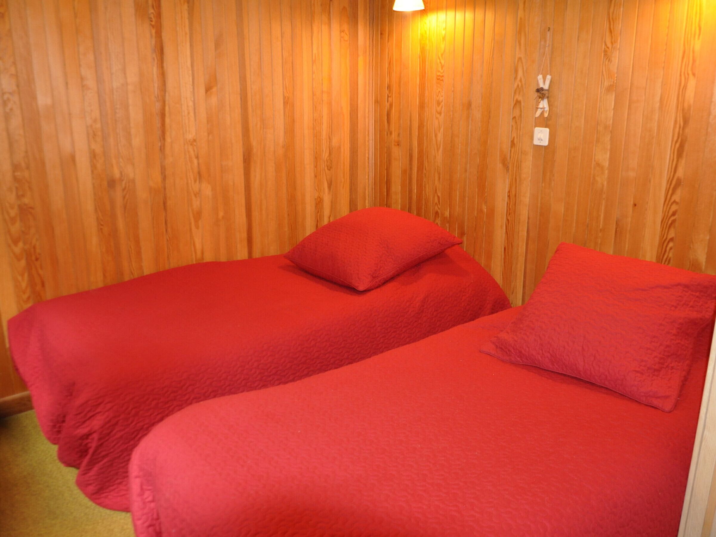 A room with two single beds, each with a red blanket and pillow. The walls are wooden paneling.