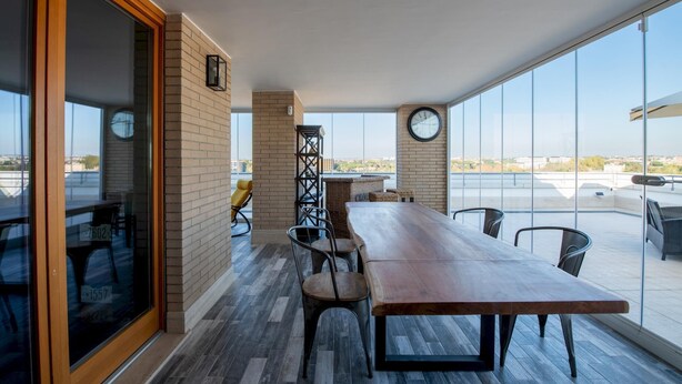 A spacious balcony with a long wooden table and chairs, a wicker chair, and a clock on the wall.