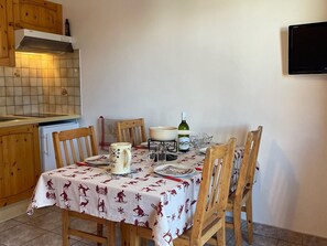 Uma sala de jantar com uma mesa posta para quatro pessoas, cadeiras de madeira, toalha de mesa branca com estampas vermelhas, uma garrafa de vinho e um jarro.