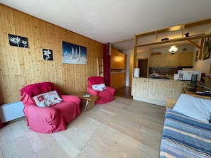 A cozy room with wooden walls, two red chairs, a small table, a bed with striped bedding, and a kitchenette area.