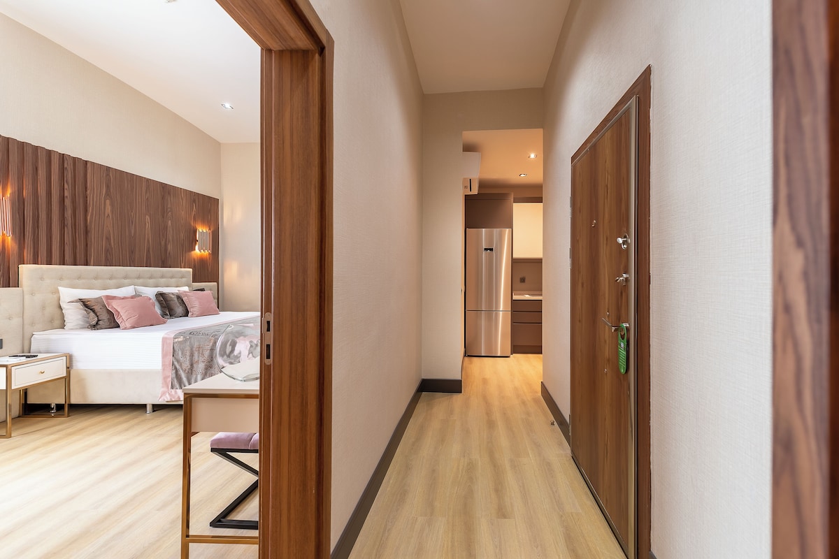 Une chambre d’hôtel moderne avec un plancher en bois, un lit doté d’une tête de lit, des tables de chevet et une vue sur la cuisine.