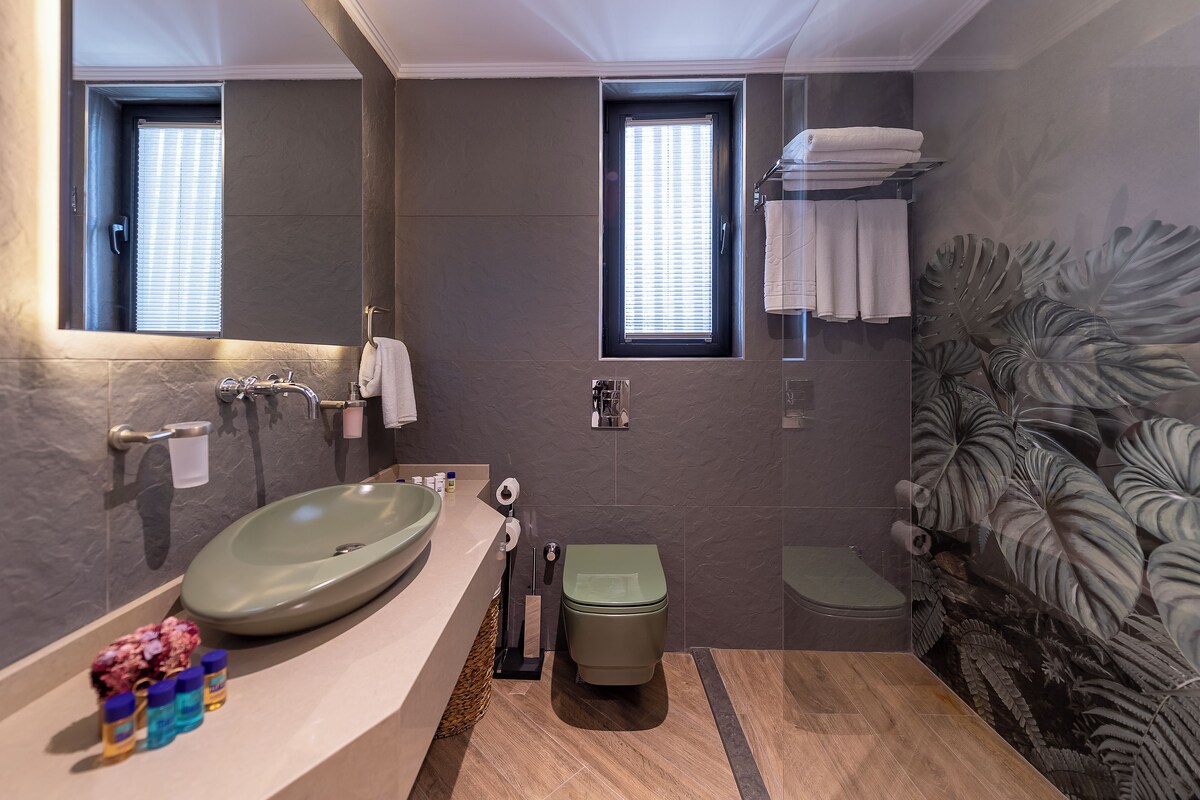 A modern bathroom with a large oval sink, a toilet, and a shower area. There are two windows with blinds, a towel rack with folded towels, and a decorative plant wall.