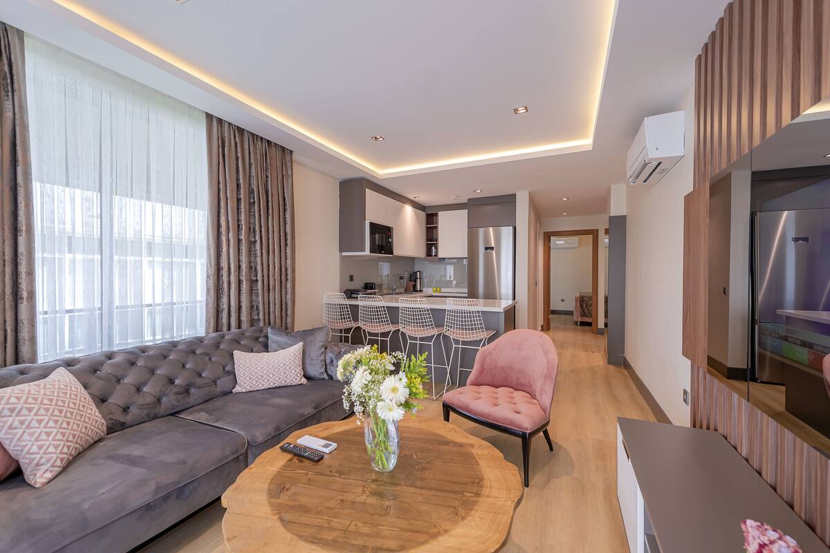 Un salon moderne avec un canapé, une table basse ronde surmontée d’un vase et un fauteuil rose. On aperçoit à l’arrière-plan une cuisine avec un réfrigérateur et un coin repas avec des chaises.