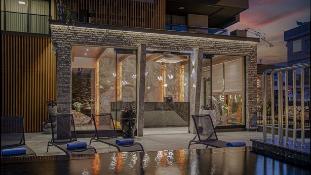 A modern hotel lobby with a pool area, featuring a stone facade, seating by the pool, and a view of the interior.