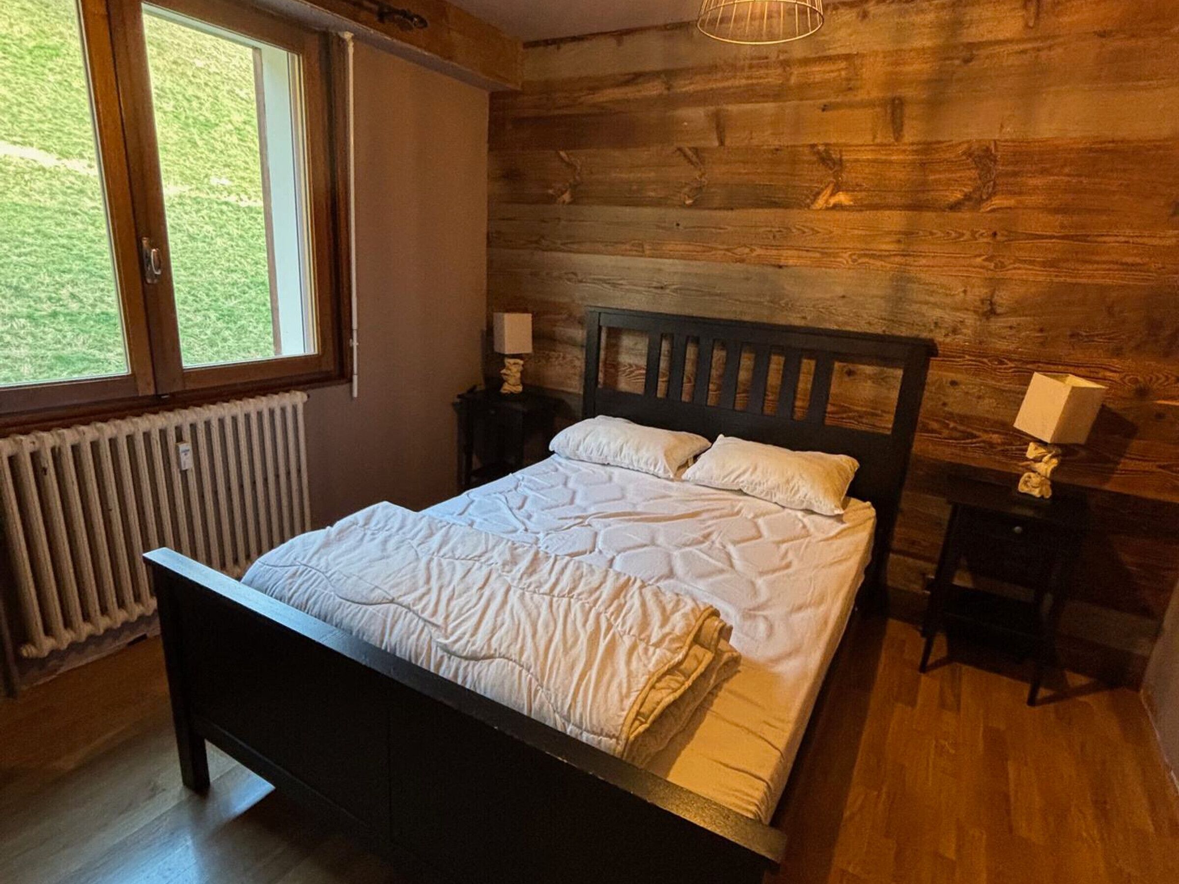 Et soveværelse med sengegavl i træ, et stort vindue, en radiator og et natbord med lampe.
