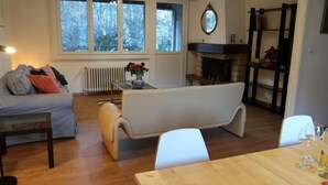 Un salon avec un canapé, une table à manger avec des verres et une assiette de nourriture, une cheminée et une vue sur les arbres à travers de grandes fenêtres.