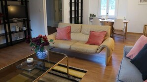 Un salon avec un canapé beige, une table basse ornée d’un vase de fleurs, un pouf à rayures et une bibliothèque.