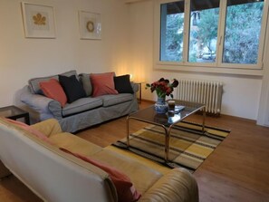 A living room with a sofa, a coffee table with a vase of flowers, and a window with a view of trees and a car.