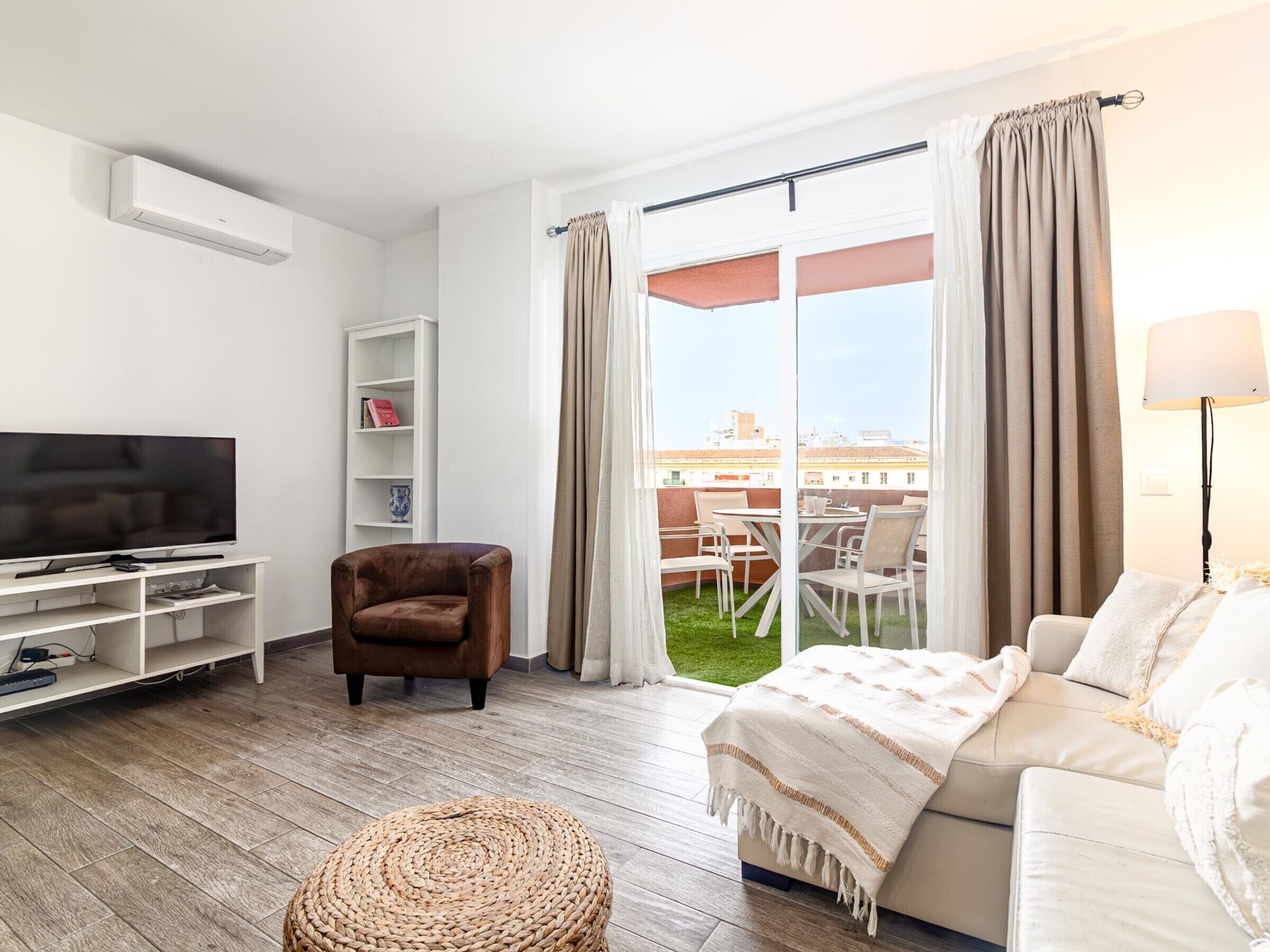 Una sala moderna con un televisor de pantalla plana, un sillón marrón, un sofá blanco y un otomano tejido. Se ve un balcón con mobiliario exterior a través de un ventanal amplio.