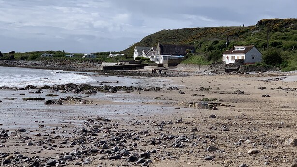 En stenet strand med hvide bygninger, heriblandt en kirke, og en bakke i baggrunden.