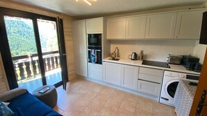 Une cuisine compacte avec des meubles blancs, un four encastré, un lave-linge et un balcon offrant une vue sur la verdure.