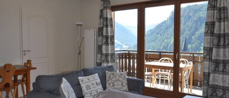 A room with a sofa, a bed, a table, and chairs. There is a view of mountains through a large window.