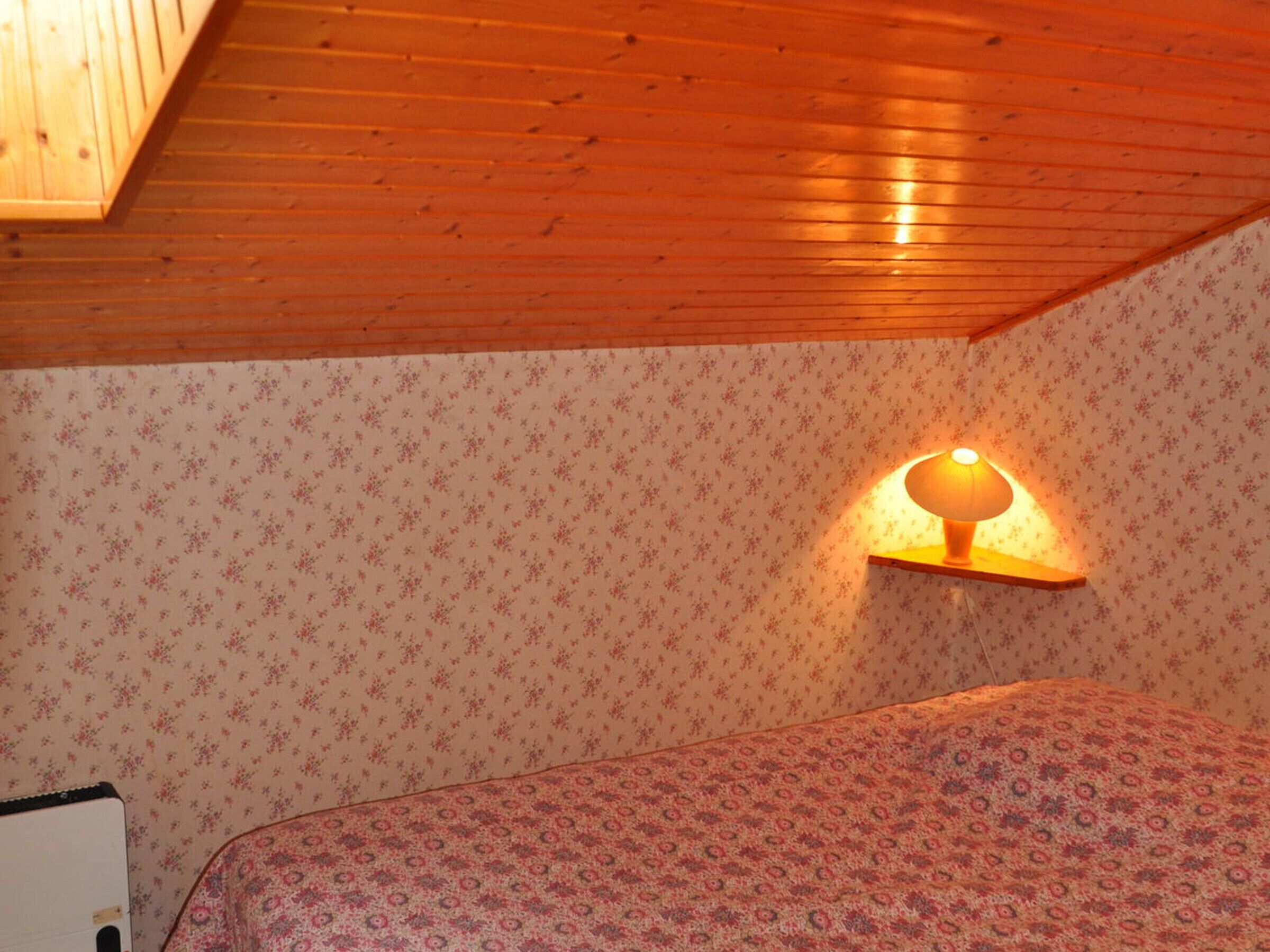 Een slaapkamer met een houten plafond, bloemenbehang, een bed met een beddekleed met dessin, en een wandlamp.