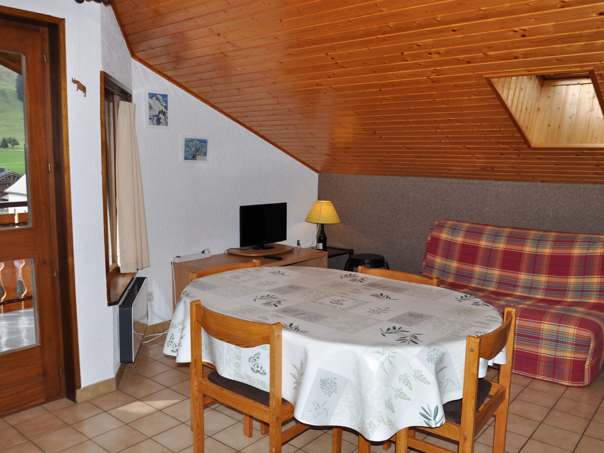 A cozy room with a wooden ceiling, a dining table with chairs, a television, and a plaid sofa.