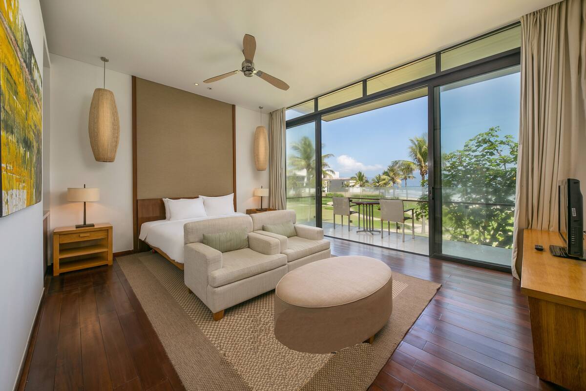 A modern hotel room with a large bed, a sofa, a round ottoman, a ceiling fan, and a balcony with a view of palm trees.