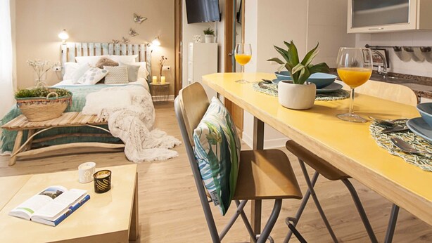 A cozy bedroom with a bed, bedside table, and a bedside lamp. There is a dining area with a table, chairs, and a potted plant. - Central Málaga Apartment: Bright, Comfortable, Fully Equipped, Near Attractions (Málaga)