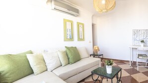 A living room with a sofa, a coffee table, and a lamp. There is an air conditioning unit on the wall and a chandelier.