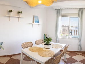 Un comedor con mesa blanca, sillas de mimbre y manteles individuales tejidos. Hay plantas en macetas sobre estantes montados en la pared y una ventana con cortinas.