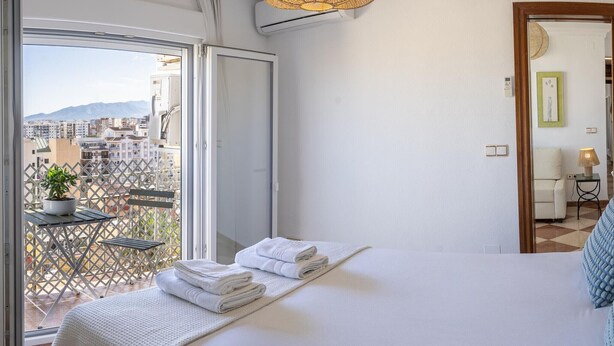 A hotel room with a bed, a balcony with a view of buildings, a hanging light fixture, and a small table with a plant.