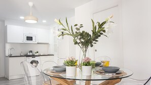 Eine moderne Küche mit weißen Schränken, einer Mikrowelle und einer Waschmaschine. Ein Esstisch mit transparenten Stühlen und eine Vase mit Blumen sind ebenfalls vorhanden.