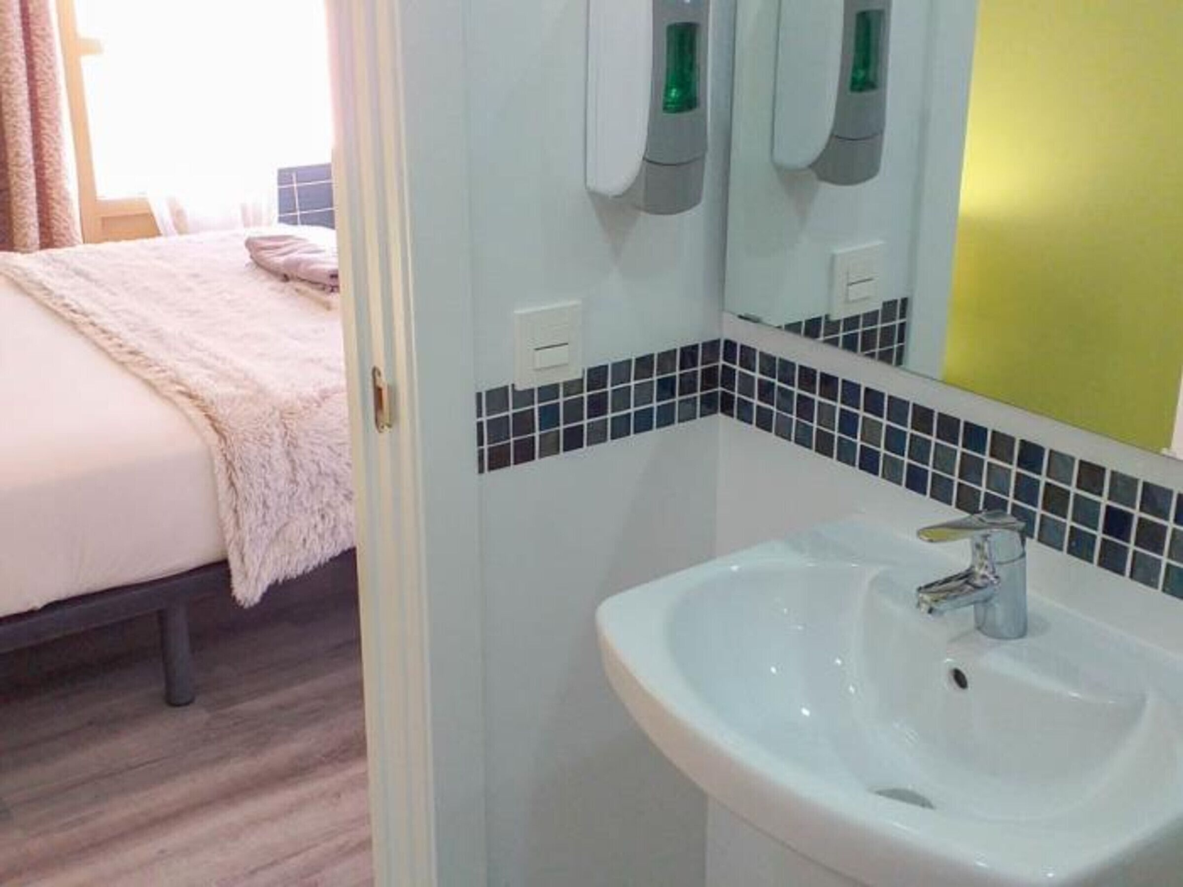A hotel bathroom with a white sink, a mirror, and a bed with a pink blanket.