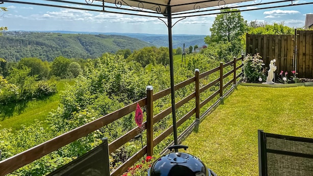 Eine überdachte Terrasse mit Blick auf ein üppiges Tal, einem Holzzaun und einer Statue.