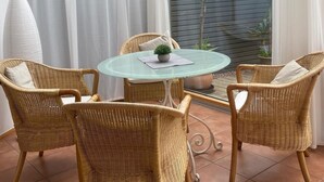 Un patio iluminado por el sol con una mesa de vidrio rodeada de sillas de mimbre, una pequeña planta en maceta y vistas de un jardín a través de un ventanal.
