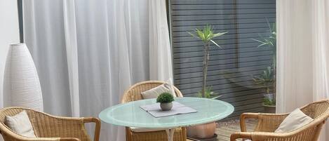 Un patio iluminado por el sol con una mesa de vidrio rodeada de sillas de mimbre, una pequeña planta en maceta y vistas de un jardín a través de un ventanal.