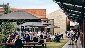 An outdoor dining area with wooden tables and black umbrellas, surrounded by a grassy lawn and a building with a stone facade. - Moonah Retreat - 4pm check out Sundays* (Fingal)