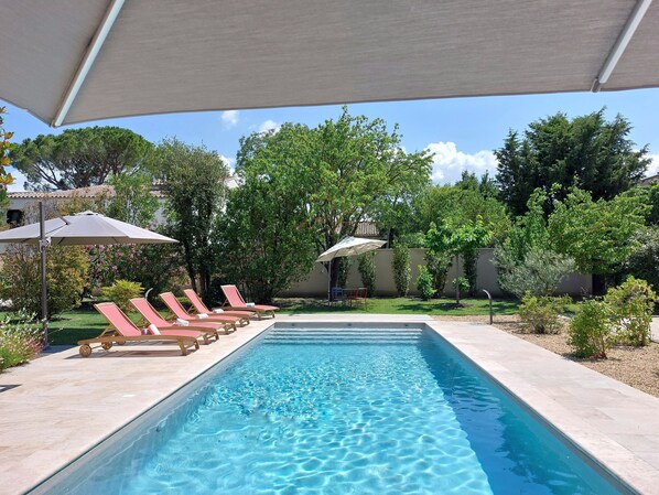 A rectangular swimming pool with clear blue water, surrounded by a tiled deck. There are several lounge chairs with red cushions and a few umbrellas providing shade.