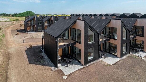 Un moderno complejo residencial con varias casas de dos pisos, con ventanas grandes y balcones, ubicado en una zona rural con un camino de tierra y vegetación escasa.