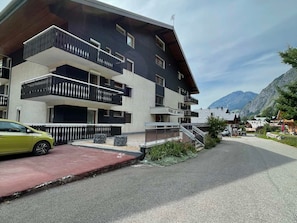 A modern multi-story building with balconies, a car parked in front, and a mountainous backdrop.