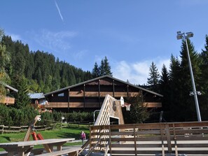 A wooden chalet with a sloped roof, surrounded by a forest. There is a basketball hoop and picnic tables in the foreground.
