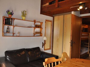 A living room with a black leather sofa, wooden shelves with books and decorative items, a wooden table, and a wardrobe with a mirror.