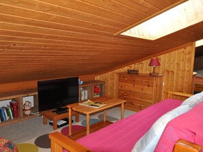 A cozy attic bedroom with wooden walls and a sloped ceiling. It includes a bed, a television, a dresser, and a small table.