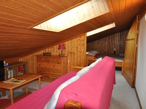 A cozy attic bedroom with a wooden ceiling, a bunk bed, a dresser, and a small table with a lamp.