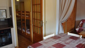 Une pièce de taille modeste comprenant un coin cuisine, un lit avec une tête de lit en bois et une table à manger recouverte d’une nappe à carreaux.
