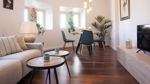 Un salon moderne comprenant un canapé, deux tables rondes, un coin repas avec des chaises, un téléviseur et un luminaire suspendu au plafond.