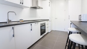 Une cuisine moderne avec des armoires blanches, un évier en acier inoxydable, un four à micro-ondes et une cuisinière. On peut voir des tabourets au comptoir et une porte au fond.