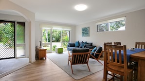 A spacious living area with a wooden floor, a sofa with cushions, a dining table with chairs, and a large window with a view of greenery.