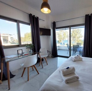 A hotel room with a bed, a desk with chairs, a TV, and large windows with curtains. - Finikas Suites & Apartments Airport (Spata-Artemida)